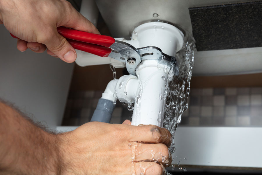 Close-up of a plumber's hands repairing a leaking sink pipe with water spraying out, showing emergency plumbing repair in action.