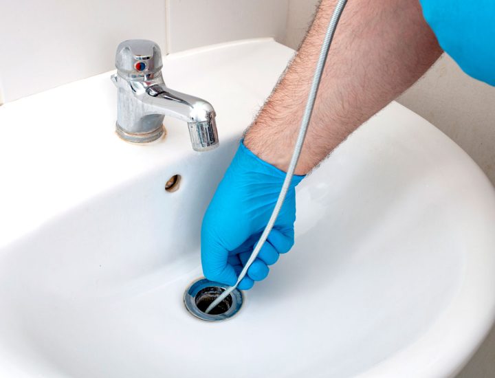 Plumber in blue gloves using a drain snake to unclog a bathroom sink, emphasizing careful and effective clog removal.