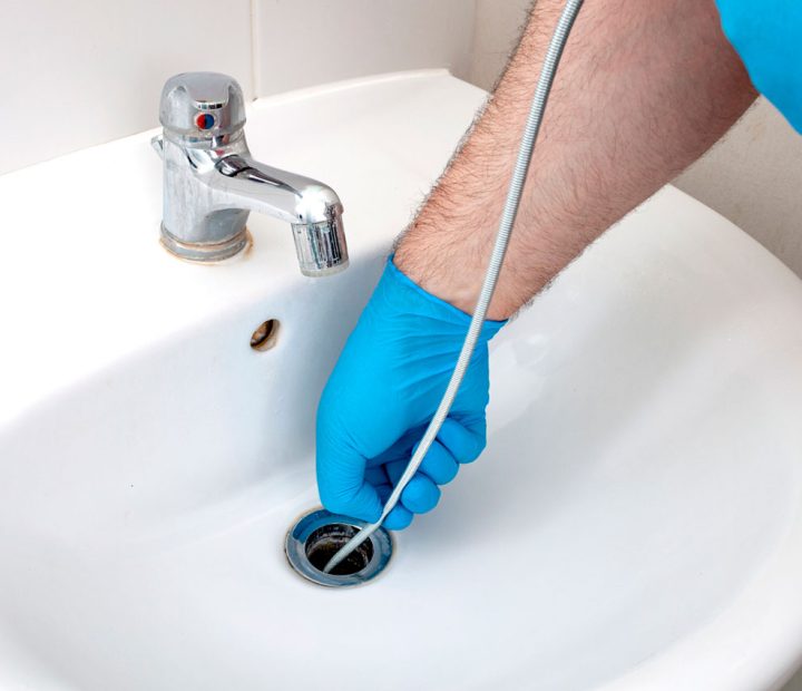 Plumber in blue gloves using a drain snake to unclog a bathroom sink, emphasizing careful and effective clog removal.