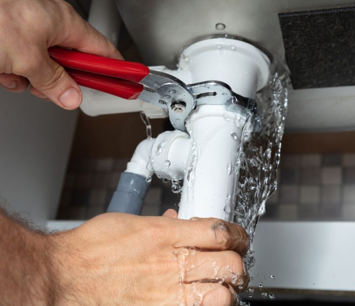 Close-up of a plumber's hands repairing a white leaking sink pipe with water splashing, illustrating emergency plumbing repair.